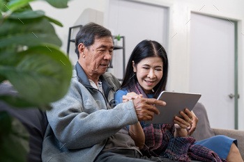 Happy elderly asian couple using tablet sit on sofa doing ecommerce shopping online on website and