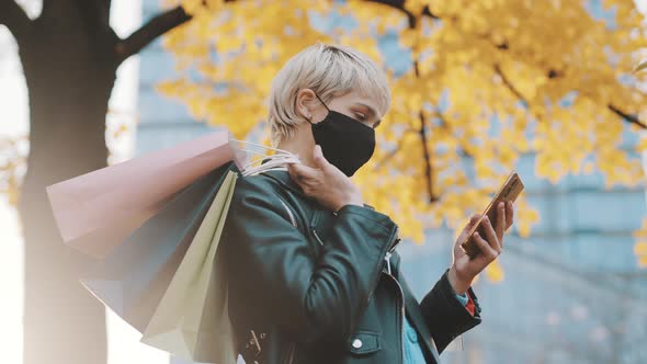 Portrait of Young Woman in Leather Jacket and Paper Shopping Bags Over the Shoulder Using Smartphone