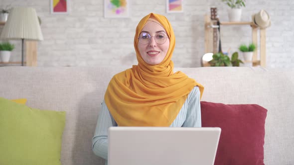 Positive Young Muslim Woman with Laptop Sitting on Sofa in Living Room at Home Looking at Camera