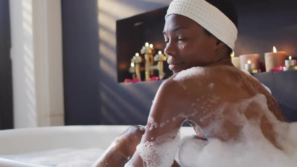 Smiling african american woman with towel taking bath and touching her skin in bathroom