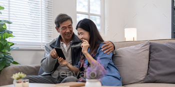 Happy elderly asian couple using smartphone sit on sofa doing ecommerce shopping online on website