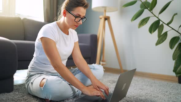 Woman with Glasses is Sitting on the Floor and Working on a Laptop