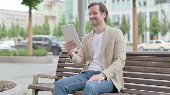 Online Video Chat on Tablet By Young Man Sitting on Bench