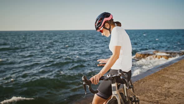 Girl in Protective Helmet Sunglasses and Sportswear is Sitting on Bike at Sea Embankment