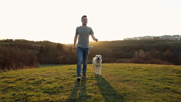 Happy Young Man Walking With His Dog On Meadow At Sunset