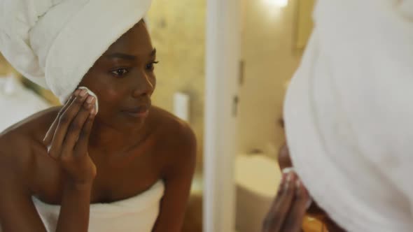 Smiling african american woman with towel watching in mirror and using pad on her face in bathroom