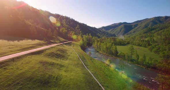 Mid Air Flight Over Fresh Mountain River and Meadow at Sunny Summer Morning. Rural Dirt Road Below.