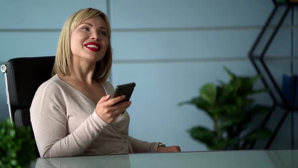 Cheerful Boss Woman Is Relaxing in Her Office, Smiling and Using Smartphone