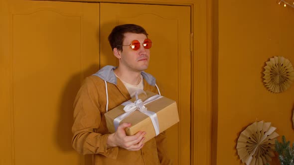 Young Man in Bright Glasses with Wrapped Gift Box Standing Near Doors in Room