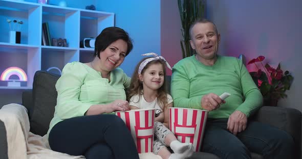 Grandfather and Grandmother With Granddaughter Sitting On Sofa At Home Watching Movie