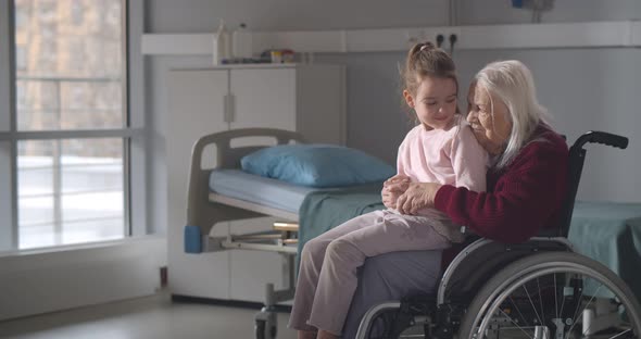 Little Girl Sitting on Lap of Old Grandmother in Wheelchair Visiting Her in Hospital Ward