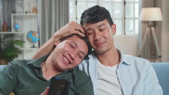 Asian Gentle Gay Couple Using Mobile Phone While Sitting On A Couch In Apartment