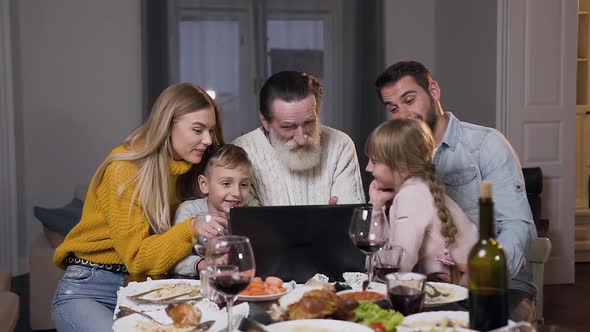 Different Generation People Sitting at the Dinner Table and Watching Funny Photos on Laptop