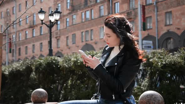 Attractive Cheerful Student Girl Sitting on Bench in Park and Chatting with a Friends