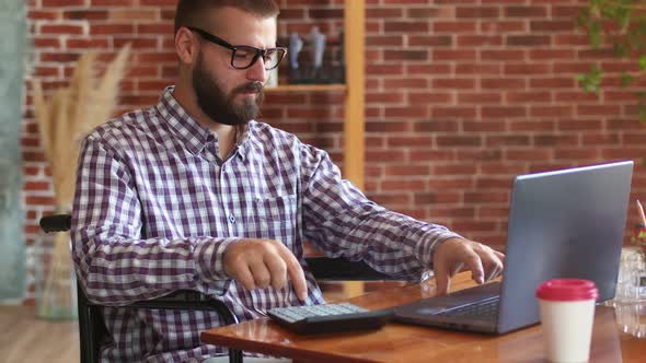 Hipster Man with Disabilities Wearing Glasses is Sitting on Wheelchair at Table with Laptop Using It