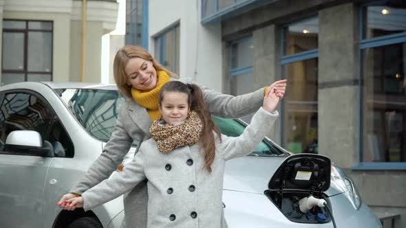 A Woman with Her Daughter Stands Near Her Electric Car and Looks at the Camera. Charging an Electric