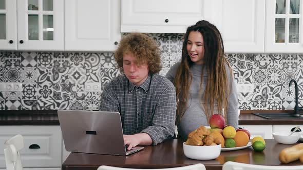 Married Man with Curls Working on Laptop in Contemporary Kitchen and Kissing His Pregnant Wife