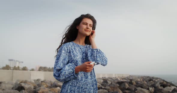 A Young Woman Smiles While Putting Headphones Into Her Ears and Looking at Sea