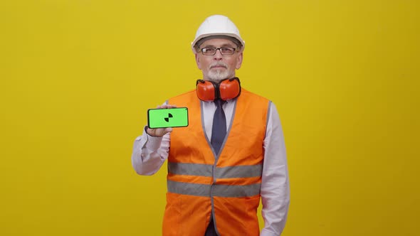 Adult man engineer in working uniform holds hand phone with greenscreen on orange background