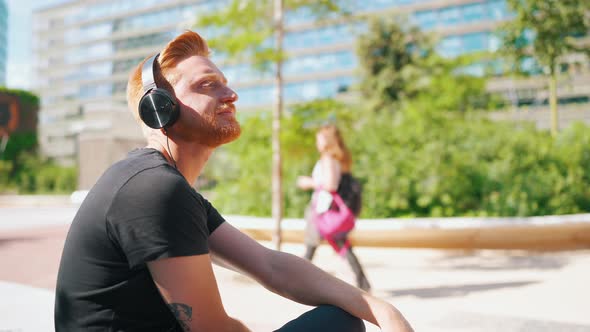 Meditative red haired man listening music in headphones while sitting on the bench