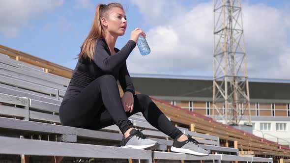 Joyful Tired Woman in Sportswear Resting and Drinking Water After Workout in Stadium