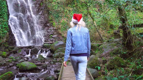 A girl in a Santa Claus hat goes to the forest to the waterfall.