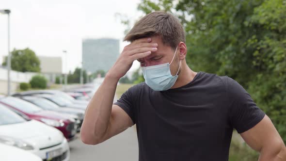 A Young Man in a Face Mask Feels Sick and Has a Headache and Coughs in a Parking Lot in Urban Area