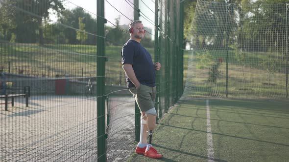 Wide Shot Portrait Happy Male Caucasian Amputee Listening to Music in Headphones Standing at Mesh