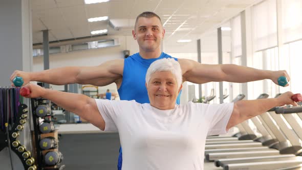 Pensioner Performs an Exercise with Dumbbells on Simulator in Gym