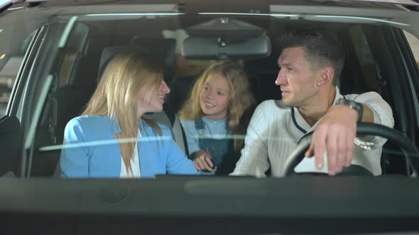 Happy Mother and Father Talking to Daughter Sitting in New Car in Dealership