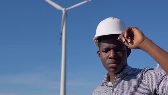 African American Electrician Engineer in a Helmet Stands Against the Backdrop of a Windmill at an