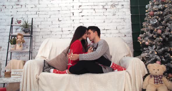 Young Happy Couple Sitting on the Sofa Near Christmas Tree with Glasses at Home. Couple Celebrating