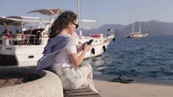 Pretty girl using smartphone while sitting at the marina. Anchored Yacht at the background