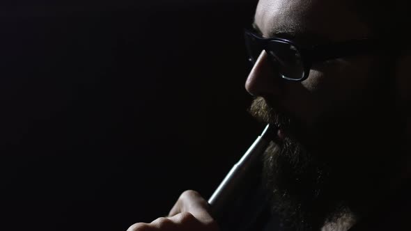 Young bearded man smokes a hookah and blow out smoke closeup on black background slowmotion