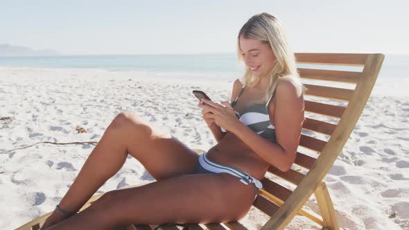 Caucasian woman sitting on a sunbed and using her smartphone on the beach