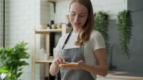 A Woman Stands in an Apron in the Kitchen and Spreads Peanut Butter on Toast