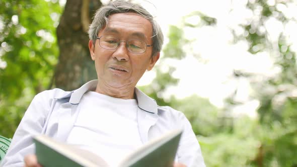 An old Asian man sits comfortably reading a book in a green park.