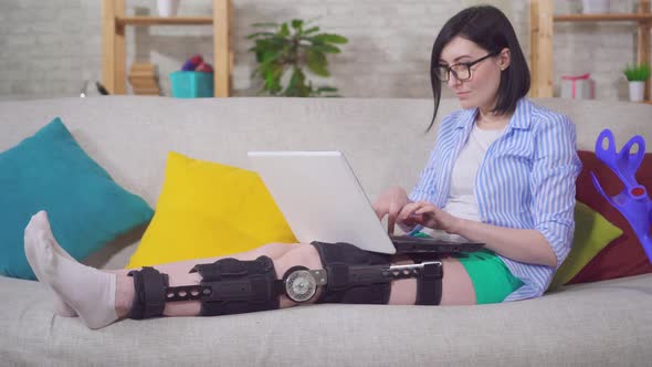 Young Woman Freelancer at Home on the Couch After Injury Knee Bend Orthosis Knee Brace with a Laptop