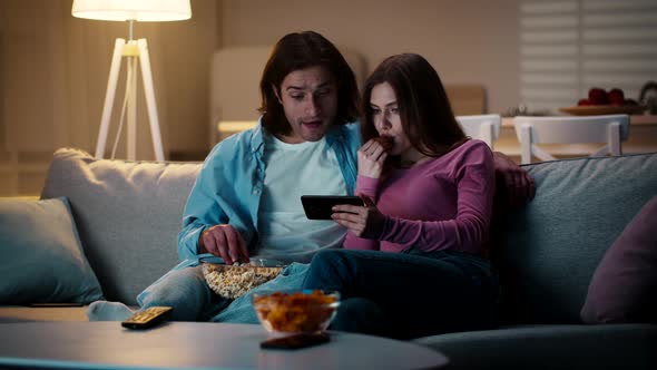Young Man and Woman Sitting on Couch in Evening and Watching Video on Smartphone Eating Popcorn and