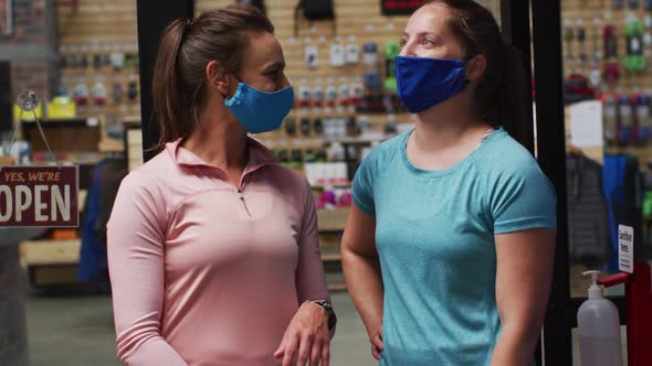 Caucasian female shopkeepers in face masks in the doorway of sports shop with arms crossed