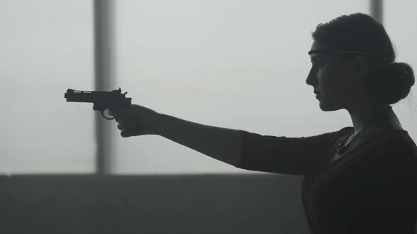 Confident Woman in Stylish Red Dress Is Aiming a Pistol in an Abandoned Dusty Building