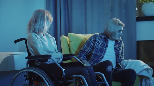 Young Man and Woman in a Wheelchair Watching TV and the Man Covering Her Legs with a Blanket