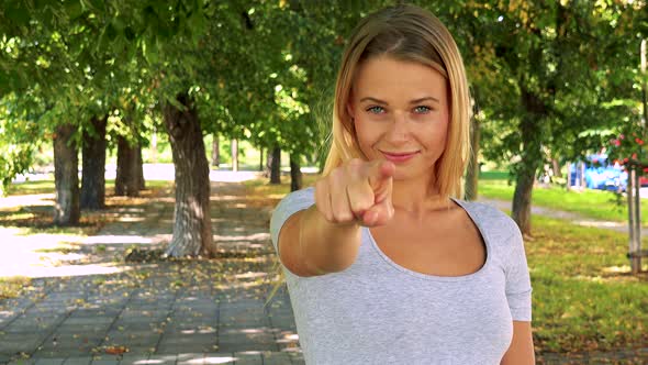 Young Pretty Blond Woman Points To Camera with Finger - Park with Trees in Background