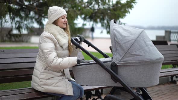 Young Mother with Newborn Child Outdoor. She Sitting on the Bench with Baby Sleeping in Pram