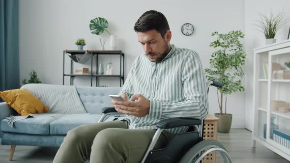 Portrait of Young Man in Wheelchair Using Smartphone Enjoying Good News Smiling Indoors at Home