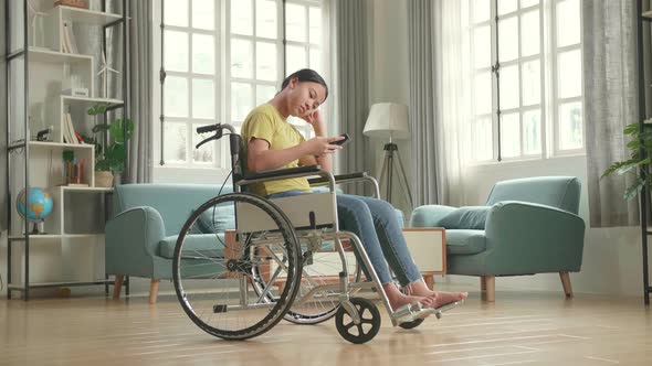 Bored Asian Kid Girl Sitting In A Wheelchair While Using Smart Phone At Home