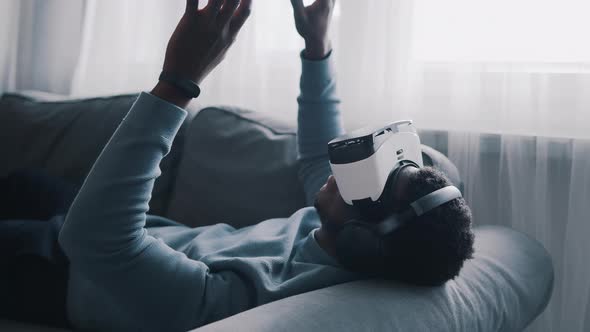 AfricanAmerican Man Wearing Virtual Reality Glasses and Headset Lying on the Sofa