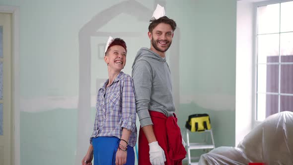 Positive Laughing Couple Posing in New House in Paper Hats Having Fun Renovating Living Room