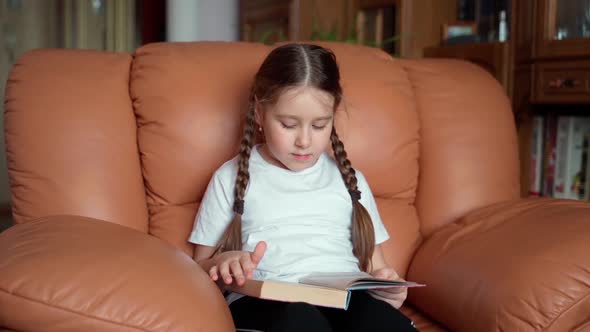 Slow motion shot of young preschool girl reading book sitting in armchair at home. Self education