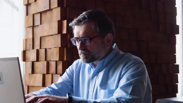 Side View of Senior Man Who Sits in Cafe in Daytime and Using the Laptop for the Remote Work